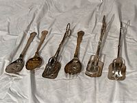 Six silver plated serving utensils arranged side by side showing their detail, shape, and size.