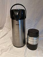 Photo showing large stainless steel coffee thermos with black plastic push-button top and handle, alongside smaller black thermos with silver ribbed band and cork lid.