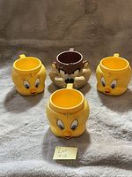 Four vintage hard plastic cups featuring Looney Tunes characters: three Tweety Birds (yellow with large blue eyes) and one Tasmanian Devil (brown with snarling face) arranged on fabric.