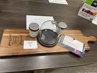 Full lot shown on wooden charcuterie board with engraved message, glass dome, candles and gift certificate/booklet on table surface.