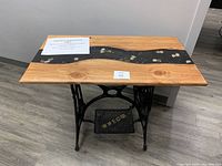 Full view of handcrafted table showing stained wood top with black epoxy river inlay containing stones and gold flakes, mounted on a vintage Williams cast iron sewing table base with treadle.