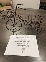 Brown metal plant holder shaped like a bicycle with two circular planters, one smaller (5.5 in) and one larger (7 in), displayed on a dark wood table with a note showing size details.