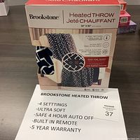 Front view of boxed Brookstone Heated Throw showing product name, features, and partial view of blue and white geometric plush throw.