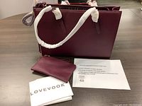 Front view of the maroon Lovevook leather-like laptop shoulder bag with detachable clutch and protective wrapping on handles, along with fabric storage bag and information sheet next to it.