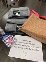 Two grey Roots Home cotton bath towels stacked with labels visible, Bath and Body Works gift card, cork-textured Bath and Body Works tote bag, and lot description paper on a wooden surface.