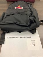 Gray unicorn backpack front with embroidered unicorn horn and flowers on zippered pocket, padded shoulder straps visible, zippered compartments with rope pulls