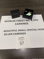 Two demi-hoop earrings with two rows of clear crystals each displayed in black box with authenticity tag and small Nic & Syd box on a printed sheet describing the earrings.