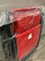 Folded red canvas zero gravity lounge chair packaged in clear plastic, showing black metal frame and elastic cords securing the fabric.