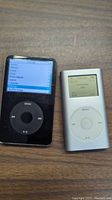 Two iPods: one black 5th generation with color screen and one white 1st generation with monochrome screen, both showing interface menus.