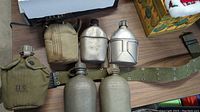 Five metal military canteens displayed on a surface; four have olive green fabric covers, one is without cover. A green military-style belt is partially visible.