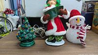 Photo showing a green decorative Christmas tree figurine with colored lights, a Santa Claus figure holding a green book, and a glittery snowman wearing a Santa hat and scarf.