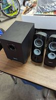 Two black satellite speakers with silver cones and one black subwoofer together on a wood surface.
