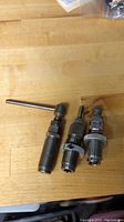 Three metal reloading reloading tools on a wooden surface including a puller, 30-30 die, and 44 Magnum sizer die
