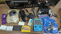 Photo showing Sega VideoNow Color handheld player, blue Game Boy Advance, multiple controllers, game cartridges, and cables on table.