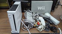 Front and side view of white Nintendo Wii console with power and reset buttons visible. Power adapter and AV cables laid beside the console.