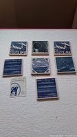 Top view of the set of seven square Delft Blue ceramic tile coasters and one certificate of authenticity laid out on a white surface, each featuring Holland America Line cruise ship or logo designs.