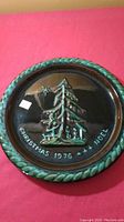Photo showing front of Christmas plate with raised tree design, text 'CHRISTMAS 1976 *** NOEL', turquoise braided border on dark brown background.