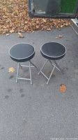 Two folding stools placed side-by-side on asphalt with dry leaves scattered in background, showing full stools and structure.
