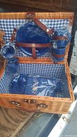 Open wicker picnic basket showing blue plastic dishes and cups secured with leather straps inside a blue and white checkered fabric lining.