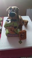 Scooby-Doo plush toy sitting on top of a green metal tin shaped as number 3 with Scooby-Doo printed on it, plus small brown plush item and Scooby-Doo collar tag charm.