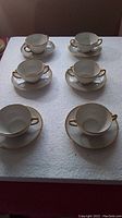 Full view of six two-handled white porcelain teacups each on a matching saucer with gold trim on edges and handles