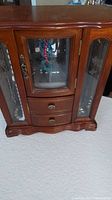 Photo of vintage wooden jewelry box showing front with glass doors and metal handles, containing various vintage jewelry pieces hung inside and sitting in drawers.