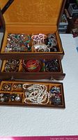 Top-down view of opened wooden jewelry box showing three drawers containing assorted vintage jewelry including bead necklaces, bracelets, earrings, and pins.