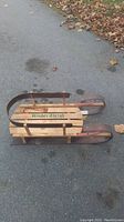 Side profile of vintage wooden child's sled showing metal runners, wooden slat seat with 'Kinder Sleigh' branding, and vertical side rails.