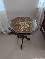 Octagonal wood side table with floral and peacock inlay on tabletop, standing upright on tripod base in carpeted room with light from window.