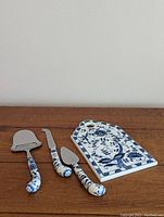 Cheese board with four matching blue and white handled cheese serving pieces arranged next to it on wooden surface, showing design and condition.