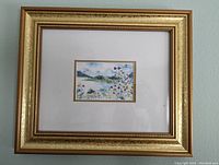 Photo of the framed watercolor painting showing the entire gold-colored frame with off-white matting and the mountain lake scene with wildflowers in the center.