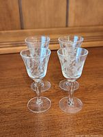 Four clear etched crystal liqueur glasses placed together on a wood surface, showing front view, decorative floral-etched design on bowl, with slender stem and circular base.