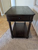 Dark wood side table showing front view with drawer and lower shelf, four turned legs, and rounded corners.