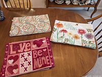 Photo of three stacks of placemats on a wooden table, showing one placemat from each set: paisley pattern, spring floral with "hello SPRING" text, and Valentine's Day with hearts and phrases.