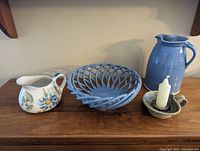 Four handcrafted pottery items arranged on wooden surface: hand painted floral pitcher, medium blue open lattice bowl, large blue pitcher, and pottery candle holder with candle.
