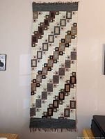 Full view of the handwoven rectangular rug or wall hanging against a wall, showing overall pattern and fringed edges.