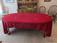 Oval red embroidered tablecloth draped over wooden table, showing detailed holiday bow and holly embroidery with cutwork pattern.