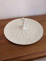 Top view of the white round ceramic serving tray showcasing its textured ornamental pattern, gold rim, and fleur-de-lis handle.