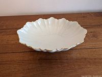 Oval white porcelain serving dish with scalloped edges and 22K gold trim, displayed on a wooden surface against a white wall.