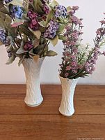 Pair of white ceramic vases with textured tree bark design, holding artificial flowers, set on wood surface against a light wall.