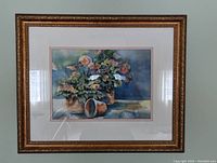 Framed watercolor painting showing a floral still life with flower pots on a table, framed under glass in a detailed gold frame.