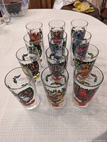 Set of 12 vintage glasses with colorful 12 Days of Christmas themed illustrations. Each shows a different day with text and images free of chips or cracks, displayed in rows on a table.