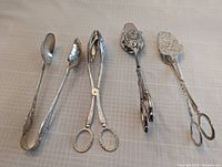 Photo showing five vintage silver-tone serving utensils including various spoons, tongs, and detailed embossed servers in good condition with some tarnishing.