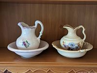 Two vintage washing bowl and pitcher sets on a wooden shelf. Left set: Peter the Potter with white and pink floral design. Right set: Haeger USA with cream base and blue floral decoration.