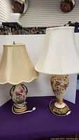 Two antique painted porcelain and brass table lamps side by side, one with beige scalloped shade and one with white bell-shaped shade on purple surface.