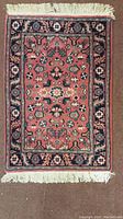 Full view of the small rectangular area rug showing central red field with intricate geometric and floral motifs, dark blue border, and white fringe tassels