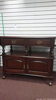 Front view of wood bar style unit showing two drawers and cupboard below, dark finish, brass handles, and wheels.