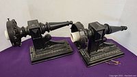 Two black cast iron wall sconces laying on a purple cloth backdrop, showing front and side views.