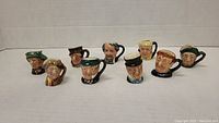 Frontal view of all nine Royal Doulton miniature Toby jugs arranged in a single row on white surface, showing detailed facial expressions and handles.