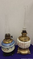 Two vintage ceramic and brass oil lamps side by side, one blue and white with floral design and the other white with pink floral design and ornate brass base, with glass chimneys on a purple surface